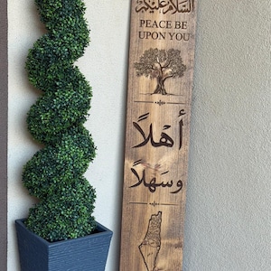 May include: A tall, rectangular wooden sign with a dark wood grain finish. It features the Arabic text 'السلام عليكم' and English text 'PEACE BE UPON YOU', an engraved olive tree, the Arabic text 'أهلاً وسهلاً', and an outline map of Palestine.