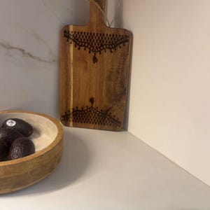 May include: A rectangular wooden cutting board with a handle and a hole for hanging, featuring dark brown pyrography patterns at the top and bottom edges. The board has a natural wood grain finish and a thin twine loop attached to the handle.