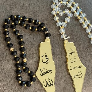 May include: Two prayer bead strands featuring faceted black and clear crystal beads with small gold-tone spacers. Each strand includes a gold-tone pendant shaped like a map, engraved with Arabic calligraphy. The black strand reads in Arabic, and the clear strand features additional Arabic text.