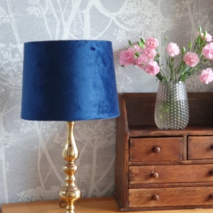 May include: A navy blue velvet lampshade sits on a gold-coloured lamp base. The lamp is on a wooden surface, with a small wooden cabinet and a vase of pink flowers in the background. A gold-framed mirror hangs on the wall.