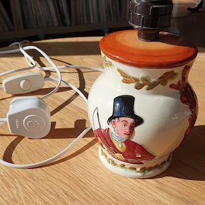 May include: A ceramic table lamp with a brown and white base featuring a hunting scene with a man in a red jacket and a black top hat. The lamp has a brown ceramic shade and a black electrical cord with a white switch.
