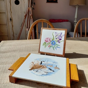 May include: Two ceramic tile trivets with wooden frames. One shows a floral design with pink, purple, and blue flowers. The other depicts Canada geese in a winter scene. Both are displayed on a wooden table.