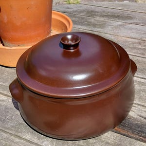 May include: A round, dark brown ceramic pot with a matching lid and two small side handles. The pot features a smooth, glossy finish and a central knob on the lid. The design is simple and functional, with a deep, rounded body and a secure-fitting lid.