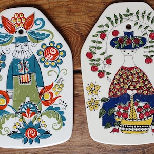 May include: Two white ceramic decorative plates with colourful folk art designs. One features a figure in blue and green clothing surrounded by flowers. The other depicts a figure with a hat and a basket of red apples.
