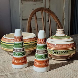 May include: A set of vintage ceramic kitchenware, including salt and pepper shakers, a bowl, and a lidded jar. The pieces have a white base with bands of orange, green, and brown. The jar is labelled "Beetroot". The items are arranged on a wooden table.