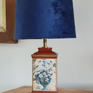 May include: A blue velvet lampshade sits atop a red and gold lamp base with a floral design. The lamp is on a wooden table.