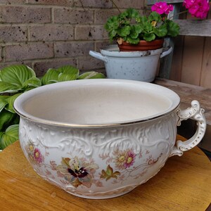 May include: A white ceramic chamber pot featuring an embossed scrollwork pattern, a gold-trimmed rim, and a decorative handle. The exterior is decorated with a colourful floral print of pink, yellow, and purple flowers with green leaves.