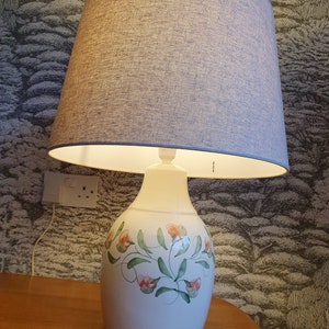 May include: A white ceramic table lamp with a floral design and a grey fabric lampshade. The lamp is turned on and illuminating the room.
