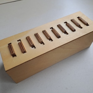 Może przedstawiać: Drewniana skrzynka z szeregiem szczelin wyciętych w górnej części. Każda szczelina ma włożony mały drewniany element.