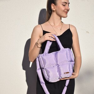 May include: A lavender quilted tote bag with a matching shoulder strap. The bag has a rectangular shape with a padded design. The handles and strap are also padded. The bag is held by a person wearing a black dress.
