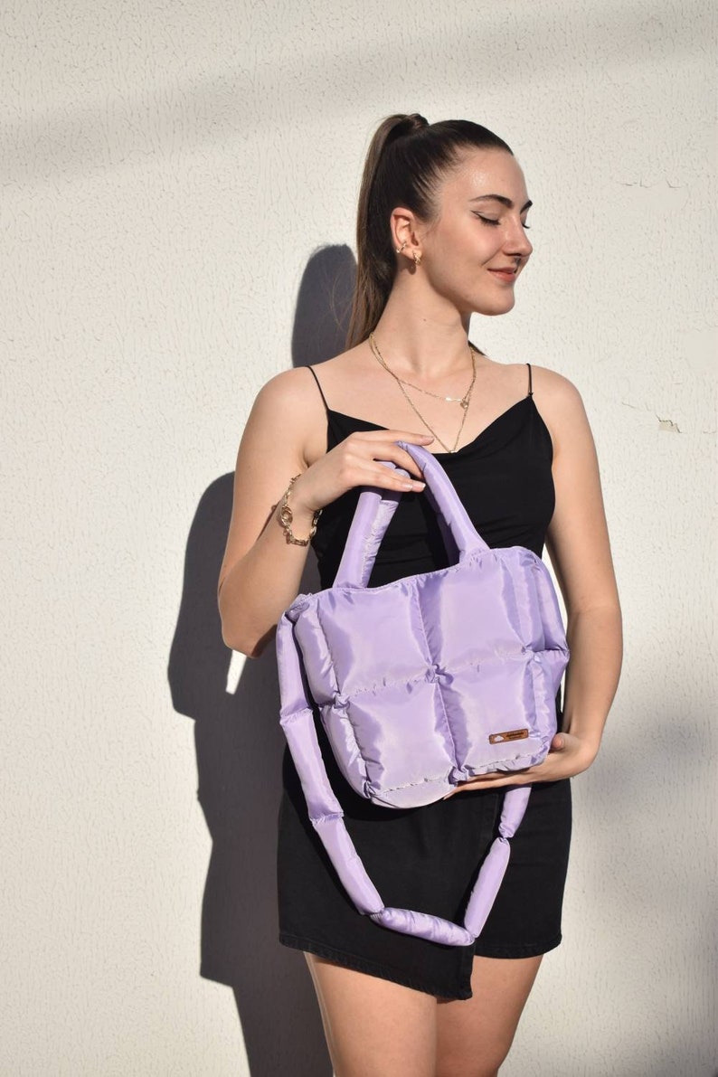 May include: A lavender quilted tote bag with a matching shoulder strap. The bag has a rectangular shape with a padded design. The handles and strap are also padded. The bag is held by a person wearing a black dress.