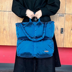 Könnte beinhalten: Eine große, gesteppte Tragetasche in leuchtendem Türkis. Die Tasche hat eine rechteckige Form mit abgerundeten Ecken und einem gepolsterten Griff. Auf der Vorderseite ist ein kleines rechteckiges Etikett angenäht. Die Tasche wird von einer Person in einem schwarzen Mantel gehalten.
