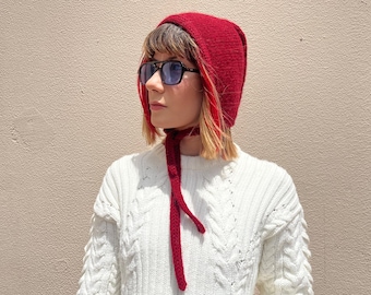 May include: A woman wearing a red knitted hat with a long tie, black sunglasses, and a white cable knit sweater.