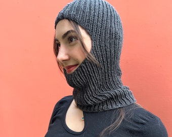 May include: A dark gray balaclava crafted from a ribbed knit fabric. The headgear features a snug fit that covers the head and neck, leaving an opening for the face. The texture of the knit is visible throughout the piece, showing vertical lines.