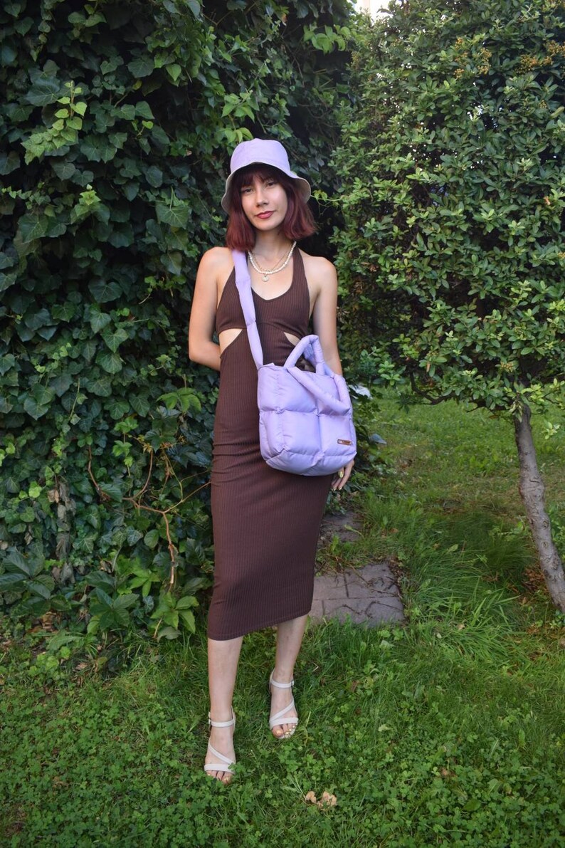 May include: A woman wearing a brown halter dress, a lavender bucket hat, and carrying a lavender quilted tote bag. She is also wearing a pearl necklace and white sandals. The background features green foliage and grass.