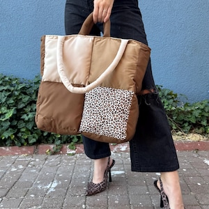 May include: A large, rectangular, quilted tote bag featuring a color-blocked design in shades of cream and brown. The bag includes two padded handles and a square front pocket with a black and white leopard print pattern on a white background.