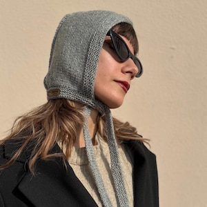 May include: A gray knitted hood with long ties, worn over the head. The hood has a textured appearance and a small, brown leather tag. The person is wearing black sunglasses and a black coat.