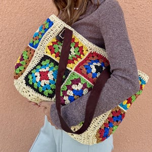 May include: A colorful crochet tote bag with a patchwork design. The bag features squares of various colors, including red, yellow, green, and blue, with a beige border and brown straps. The bag is held by a person.
