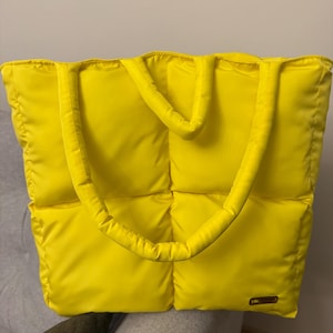 Peut inclure: Un sac fourre-tout carré, jaune vif, matelassé avec une texture rembourrée et gonflée. Le sac présente deux épaisses bretelles assorties de couleur jaune et une petite étiquette rectangulaire de marque fixée en bas à droite du panneau avant.