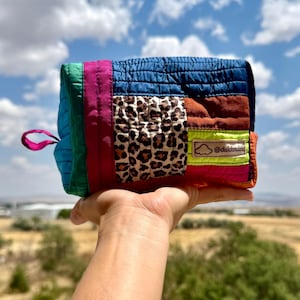 May include: A rectangular quilted pouch featuring a patchwork design of blue, green, magenta, brown, and neon yellow fabric panels with a leopard print section. A small pink loop is on the side, and a label displays a cloud icon and @daldandalo.