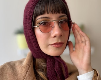 May include: A woman wearing a maroon knitted hat with a long scarf tied around her neck. She is wearing pink tinted aviator sunglasses and a brown jacket.