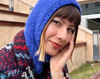 May include: A vibrant blue knitted hood, worn over a patterned coat. The hood is a solid, bright blue color, and the coat features a mix of red, black, and blue patterns. The image is a close-up shot.