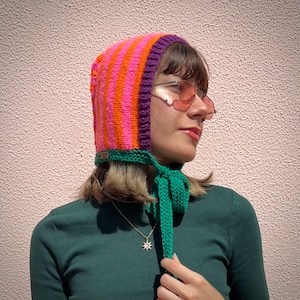Peut inclure: Un bonnet en tricot rayé rose et orange avec une cravate verte. Le bonnet a une bordure violette et est porté par une personne aux cheveux bruns.