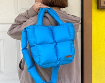 Könnte beinhalten: Eine hellblaue gesteppte Tragetasche mit einem bauschigen Design und einem braunen Lederetikett. Die Tasche hat einen einzigen oberen Griff und einen langen Schultergurt.