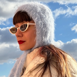 May include: A person wearing a light blue fuzzy hood and scarf, with cream-coloured sunglasses. The person is also wearing a cream-coloured jacket. The background shows a blue sky with white clouds.