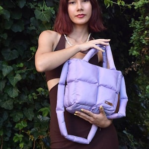 May include: A lavender quilted tote bag with matching handles. The bag has a rectangular shape with a padded design. The bag is held in front of a green leafy background. The bag has a small brown label.