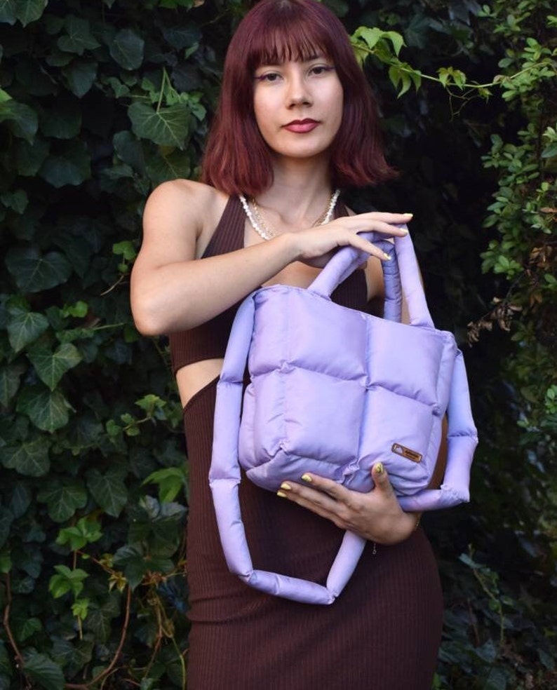 May include: A lavender quilted tote bag with matching handles. The bag has a rectangular shape with a padded design. The bag is held in front of a green leafy background. The bag has a small brown label.