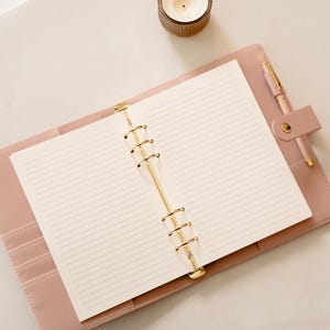 May include: A pink leather-bound planner, open to reveal lined paper and gold-coloured metal rings. A matching pink pen is secured in a loop. A small candle in a decorative holder is in the background.