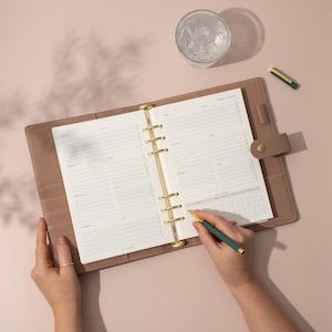 May include: A brown leather bound planner with gold rings and a gold pen. The planner is open to a weekly spread with lined pages for writing appointments and tasks. The planner is on a light pink surface.