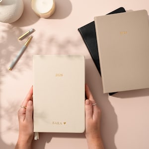 May include: A cream-coloured 2026 planner is held open, with the name "SARA" and a heart symbol printed on the cover. A gold pen and two other planners are also visible. The planner is on a light pink surface.