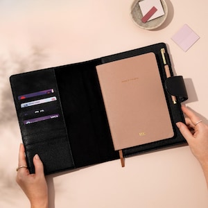 May include: Black leather planner with a blush-colored notebook inside. The notebook has gold lettering and a matching pen. The planner has card slots and a magnetic closure. The planner is open, revealing the interior.