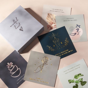 May include: A set of affirmation cards with gold foil lettering on a light blue background. The cards feature various designs, including a hand holding a rose, a person meditating, and a plant. The box the cards are in is light blue with the words "mindful heart" in gold foil lettering.