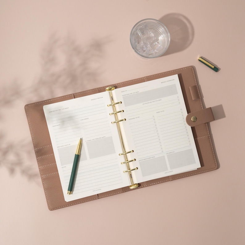 May include: A brown leather bound planner with gold rings and a green pen. The planner is open to a weekly spread with the words "Weekly Notes" at the top of the page.