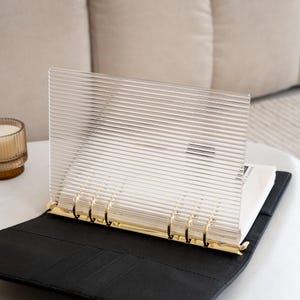 Op de afbeelding: Een zwarte lederen planner met gouden ringen en een transparante plastic hoes met een geribbelde textuur.