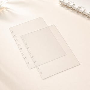 May include: Transparent plastic binder dividers with pre-punched holes for ring binders. Two dividers are visible, one slightly smaller than the other, overlapping on a light beige surface. The dividers are clear and appear to be made of a thin, flexible material.