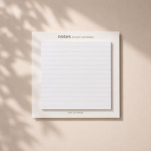 Könnte beinhalten: Ein quadratischer, weißer Haftnotizblock mit horizontalen Linien, der auf einer größeren weißen Trägerkarte befestigt ist. Der Text notes STICKY NOTEPAD ist oben und FAYE CO PAPIER unten auf der Trägerkarte in schwarzer Schrift gedruckt.