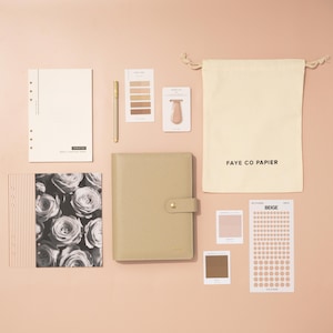 Può includere: Un assortimento di articoli di cancelleria, tra cui un'agenda beige, una penna, adesivi e un sacchetto con coulisse con la scritta "FAYE CO PAPIER". L'immagine presenta anche un taccuino con stampa floreale e campioni di colore, il tutto disposto su una superficie rosa.