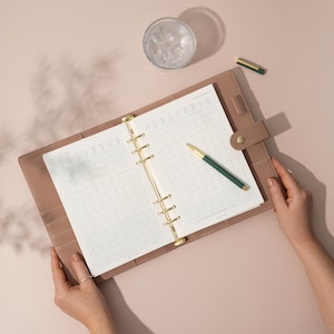 May include: A light brown leather bound planner with gold rings and a green pen lying on top of the open pages. The planner is open to a weekly grid layout.