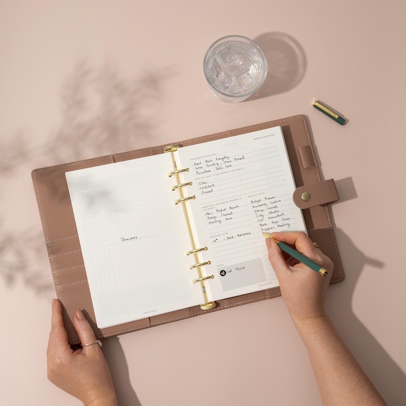 May include: A brown leather bound planner with gold rings and a white page open to a monthly calendar view. The page has handwritten notes and a to-do list. The planner is on a light pink surface.