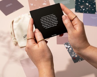 Puede incluir: Una persona sostiene una tarjeta negra con texto blanco, rodeada de otras tarjetas con diseños florales y abstractos. La tarjeta dice: "Honra el ritmo de mi vida... Estoy destinado a ser." Una pequeña bolsa con cordón es visible.