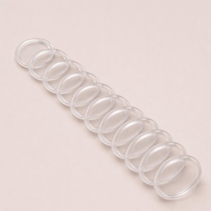 Puede incluir: Una serie de anillos de plástico transparentes de forma ovalada, dispuestos en diagonal sobre un fondo rosa claro. Los anillos transparentes están espaciados uniformemente, posiblemente para manualidades u organización.