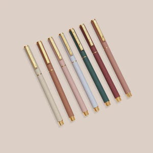 Könnte beinhalten: Sieben farbige Stifte mit goldfarbenen Akzenten, diagonal auf einer hellbeigen Oberfläche angeordnet. Die Stifte sind in den Farbtönen Creme, Braun, Rosa, Hellblau, Grün, Burgunderrot und Pfirsich gehalten. Die Stiftspitzen und -oberteile sind goldfarben.