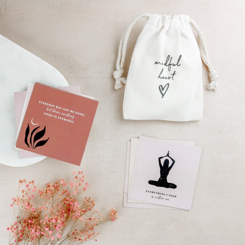 May include: A set of affirmation cards with a pink and white color scheme. The cards feature inspirational quotes and illustrations. The cards are packaged in a white drawstring bag with the text "mindful heart" and a heart symbol.