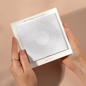 May include: A white habit tracker sticky notepad with a grid design, held by hands. The notepad has the text "HABIT TRACKER STICKY NOTEPAD" at the top and "FAYE CO PAPIER" at the bottom. The grid is a semi-circular design.