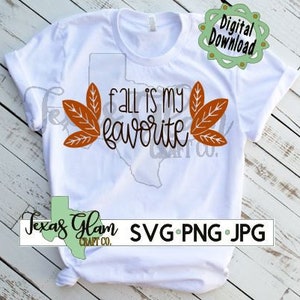 Puede incluir: Camiseta blanca con el texto "fall is my favorite" en letras marrones y dos diseños de hojas naranjas a cada lado del texto. El texto está en una fuente cursiva. La camiseta está sobre una superficie de madera.