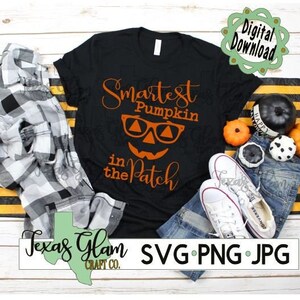 Puede incluir: Camiseta negra con una calabaza con gafas y el texto "Smartest Pumpkin in the Patch". La camiseta está sobre una mesa con vaqueros, una camisa de cuadros y calabazas.